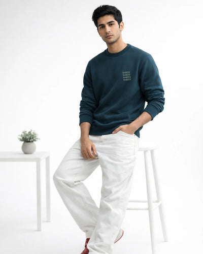Man wearing a dark blue sweatshirt and white pants sitting on a white stool against a white background.