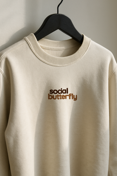 Beige sweatshirt with 'social butterfly' text on a hanger against a neutral background